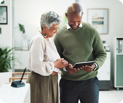 An older woman and a younger man smile while looking at a tablet together in an office