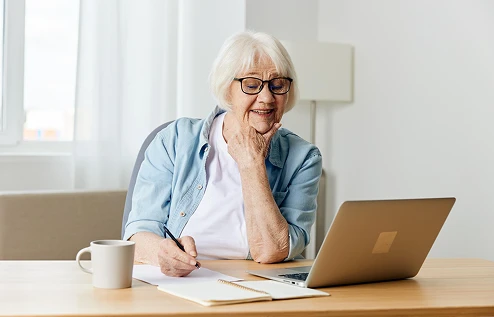An elderly woman with glasses smiles while using a laptop at a table