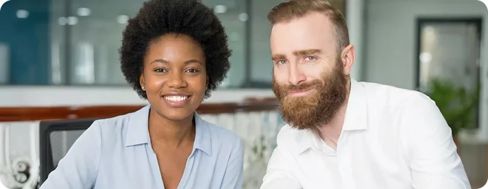 Smiling man and woman sitting together in a bright office. The woman wears a light blue shirt, and the man has a white shirt and beard. Warm, friendly atmosphere.