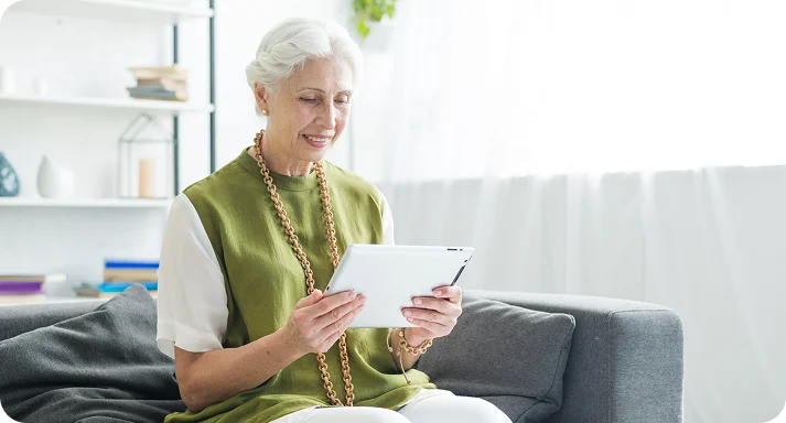 Elderly woman with white hair sits on a gray couch, smiling while using a tablet. Sunlit room with a white curtain and shelves in the background conveys a relaxed mood.