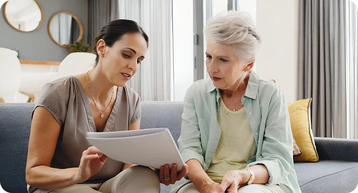 Two women sit on a sofa discussing documents. The woman on the left, in a taupe blouse, points at papers. The older woman, in a mint cardigan, listens attentively.