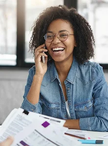 A woman wearing glasses and a denim shirt smiles while talking on a phone indoors. Papers are spread on a table in front of her. The atmosphere is cheerful.