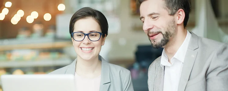 Two people in business attire, a woman and a man, smile while looking at a laptop in a warmly lit café, conveying a collaborative and positive atmosphere.