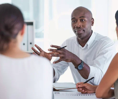 A man in a white shirt discusses ideas with two colleagues at a meeting. He gestures while holding a pen, conveying a focused and professional atmosphere.