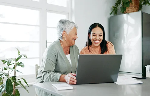 Two women smile while looking at a laptop. They sit at a kitchen counter, with natural light flooding in. A plant and fridge are in the background.