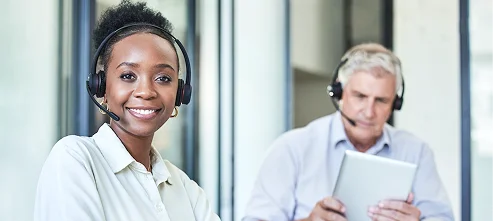 A smiling woman with a headset in an office, focusing on the camera. Behind, an older man with a headset concentrates on a digital tablet. Bright, professional setting.