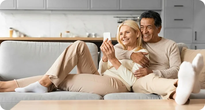 A smiling couple sits on a gray sofa in a cozy living room. The woman holds a smartphone while the man embraces her from behind. They both wear beige outfits.